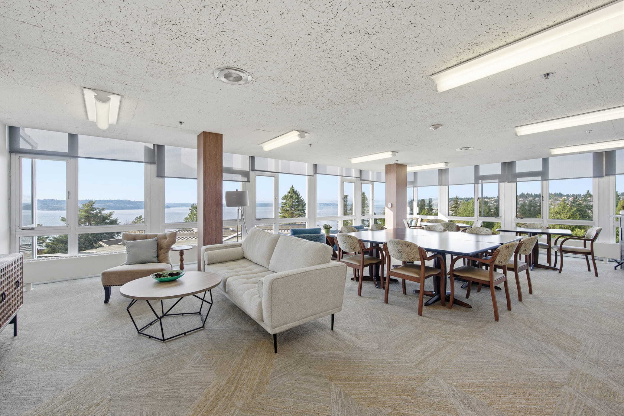 A bright community lounge at Judson Park with comfortable seating, large windows, and views of the surrounding landscape and water.