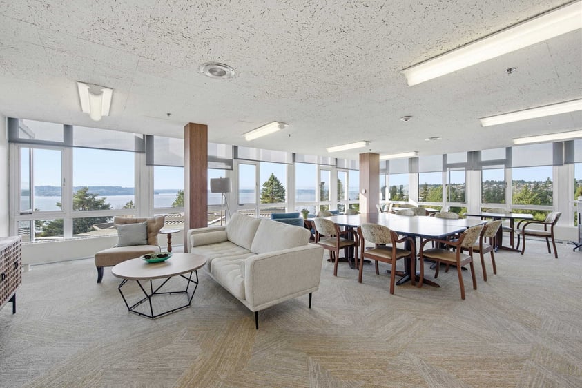 A bright community lounge at Judson Park with comfortable seating, large windows, and views of the surrounding landscape and water.