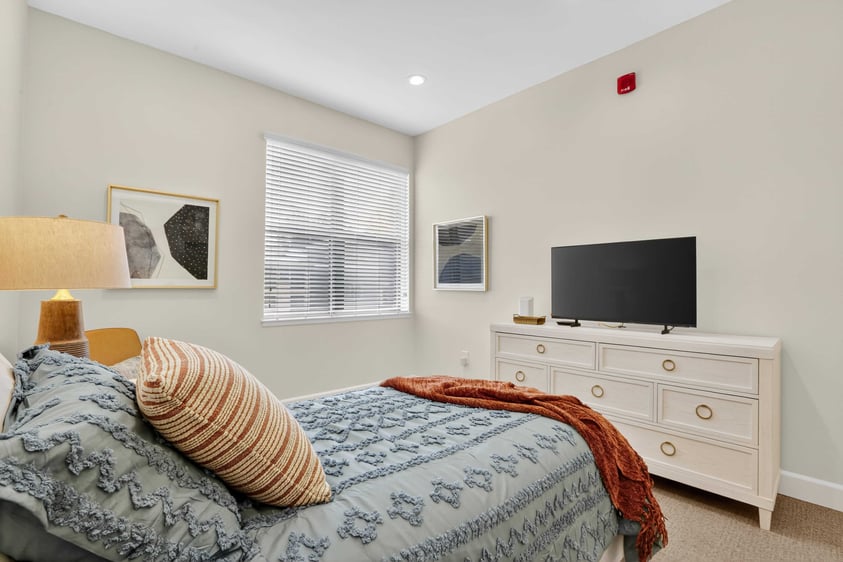 Bedroom in an assisted living apartment at The Terraces of Los Gatos with a bed, dresser, and natural light.