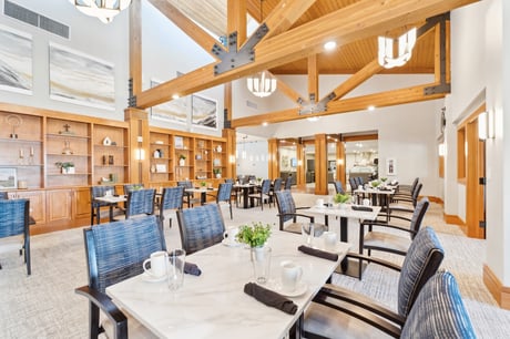 A well-lit dining area with wooden beams, blue upholstered chairs, and tables set with dining ware and decorative plants.