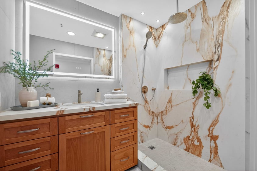 Modern bathroom with a walk-in shower, marble-style walls, wood vanity, and lighted mirror.