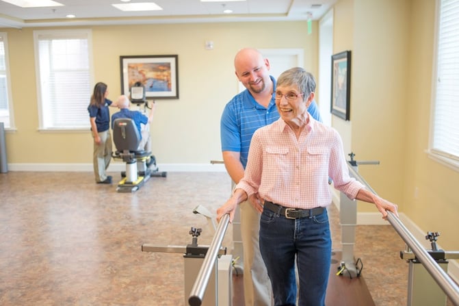 A physical therapist directs a woman walking through parallel bars.