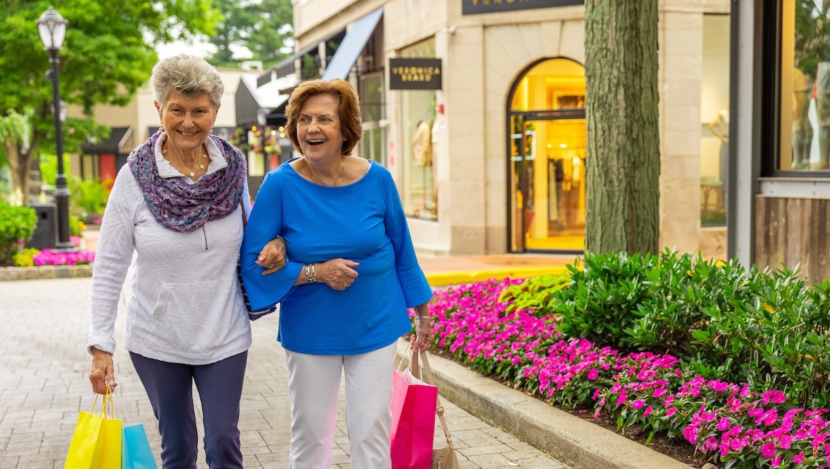 Two senior women with linked arms holding shopping bags and walking in an outdoor shopping area