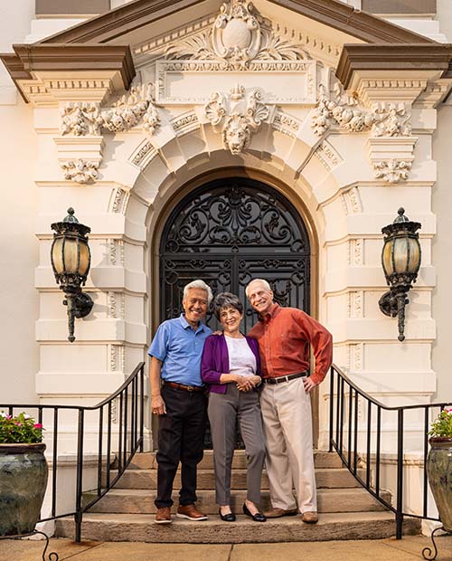 Three senior friends smiling on the steps of a mansion