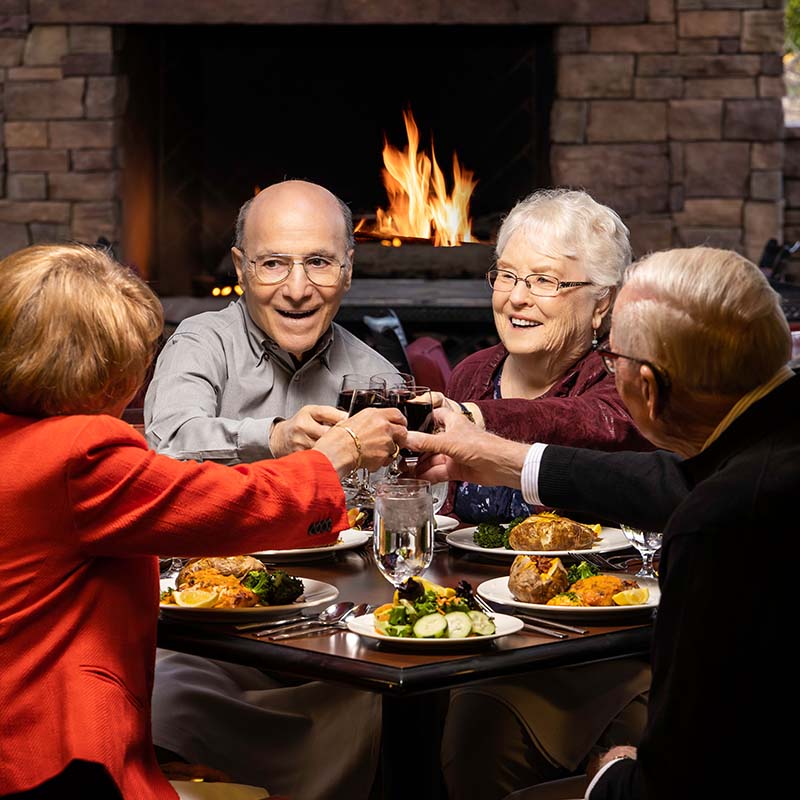 Four senior friends toasting with wine glasses and eating dinner on an outdoor patio with a fireplace