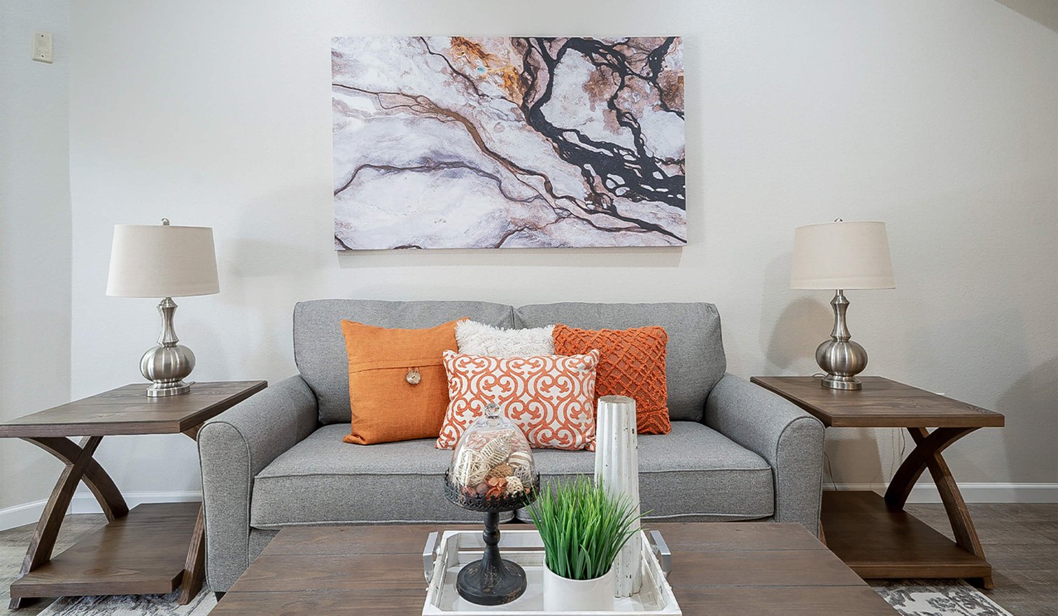 Living room with orange accent pillows on a gray couch, two end tables with lamps, a coffee table and a painting