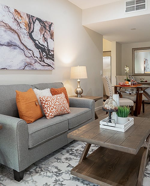 Living room with orange accent pillows on a gray couch, end table with lamp, coffee table and a painting