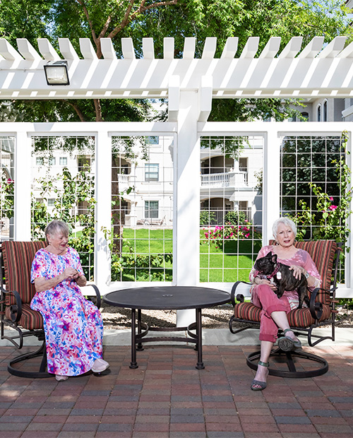 Two senior women, one holding a small black dog, sitting under a pergola at The Terraces of Phoenix