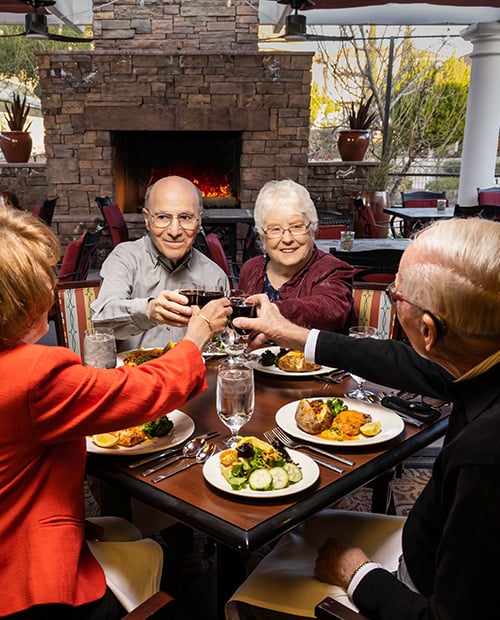 Four senior friends toasting with wine glasses and eating dinner on an outdoor patio with a fireplace