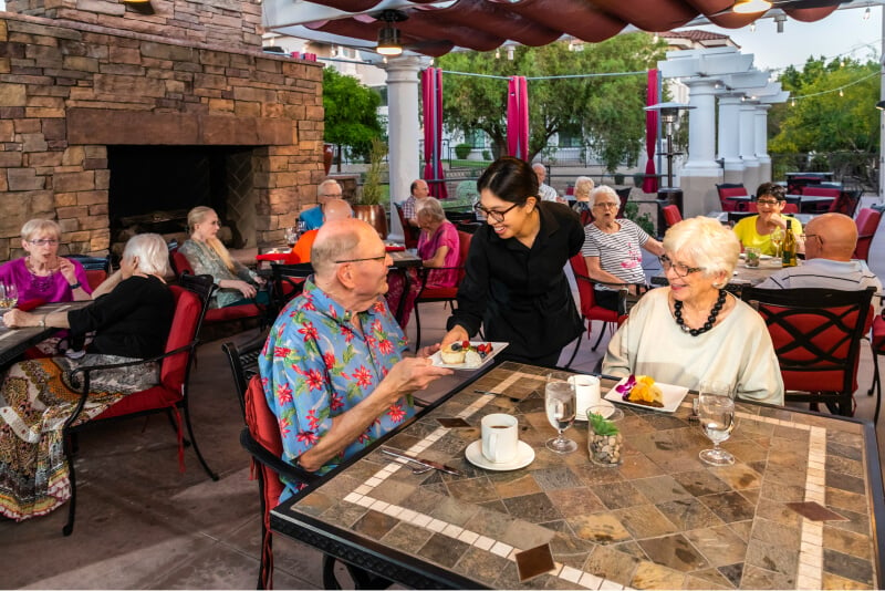 Female server handing a plate of food to a senior man eating with a senior woman on a patio