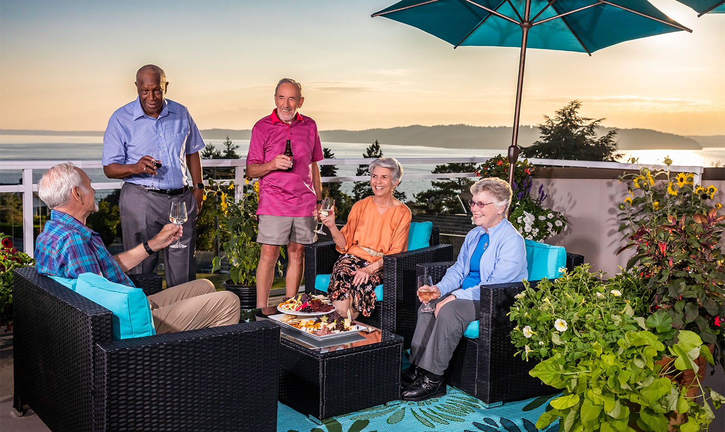 Group of five senior friends enjoying food and drinks on a balcony overlooking the Puget Sound