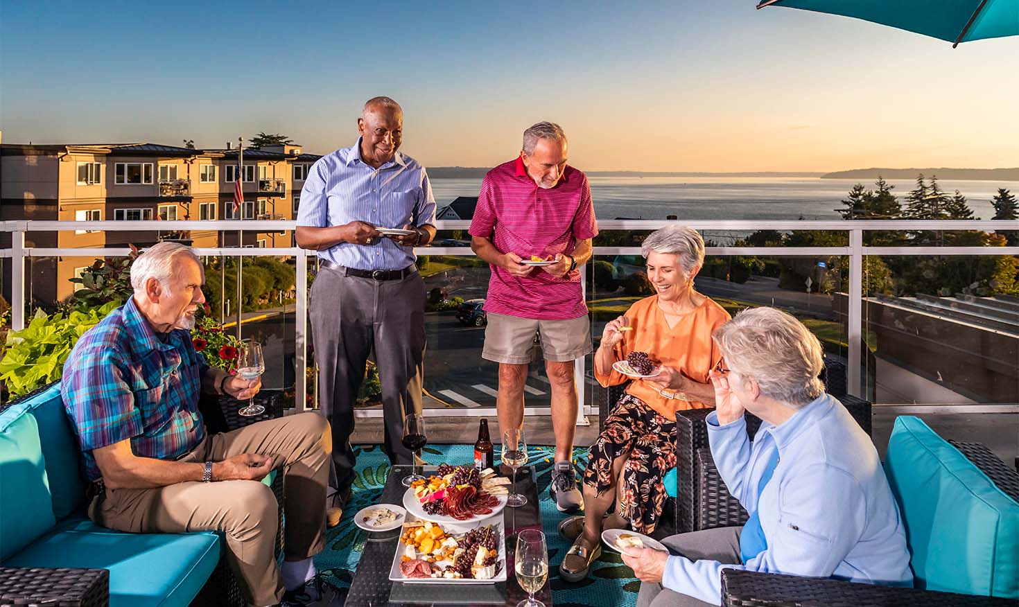 Group of five senior friends enjoying food and drinks on a balcony overlooking the Puget Sound