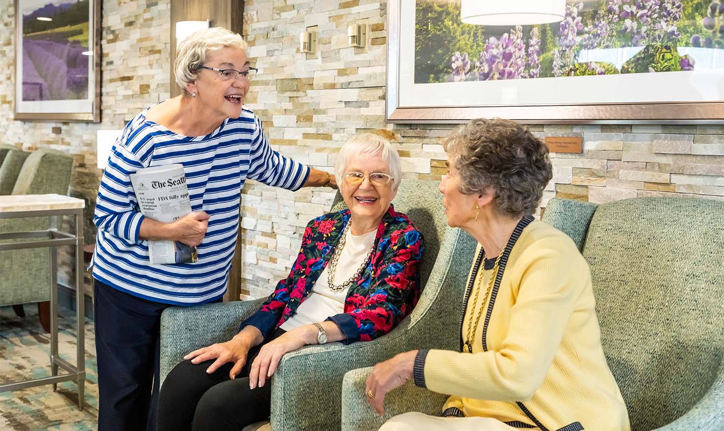 Three senior women, two sitting, one standing and holding a newspaper, chatting