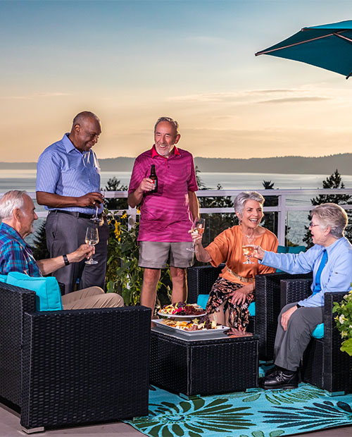 Group of five senior friends enjoying food and drinks on a balcony overlooking the Puget Sound