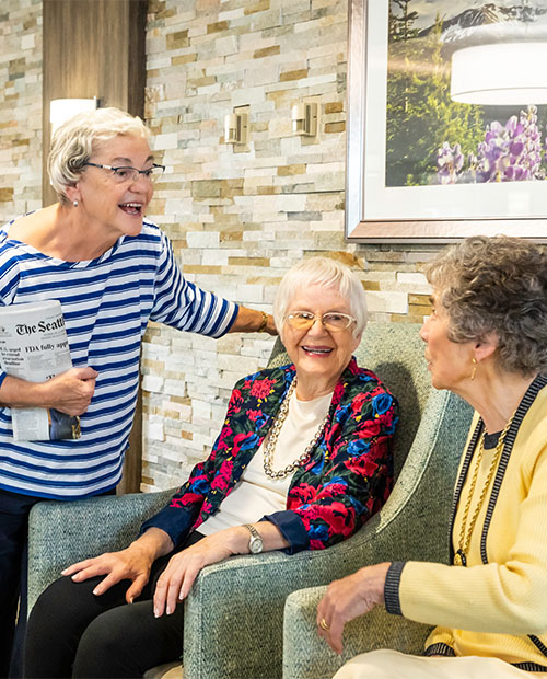 Three senior women, two sitting, one standing and holding a newspaper, chatting