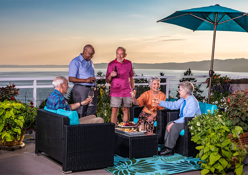 Group of five senior friends enjoying food and drinks on a balcony overlooking the Puget Sound