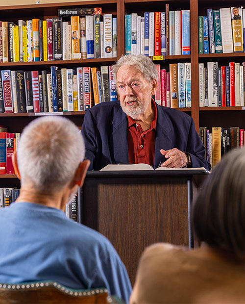 Senior man at a podium in front of bookshelves lecturing to crowd
