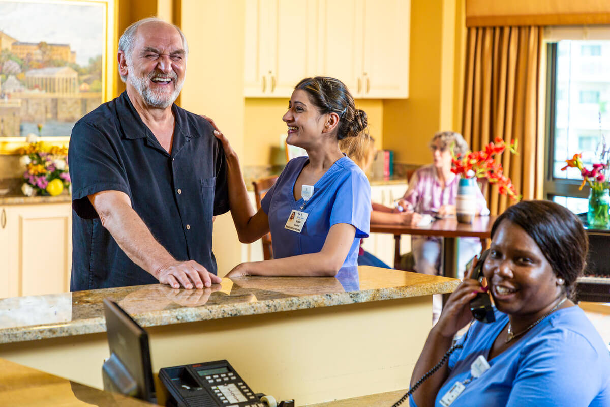 Smiling senior man and female nurse standing at nurse's station as another nurse talks on the phone