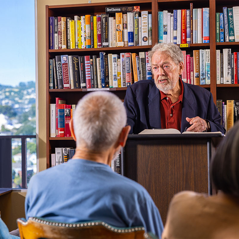 Senior man at a podium in front of bookshelves lecturing to crowd