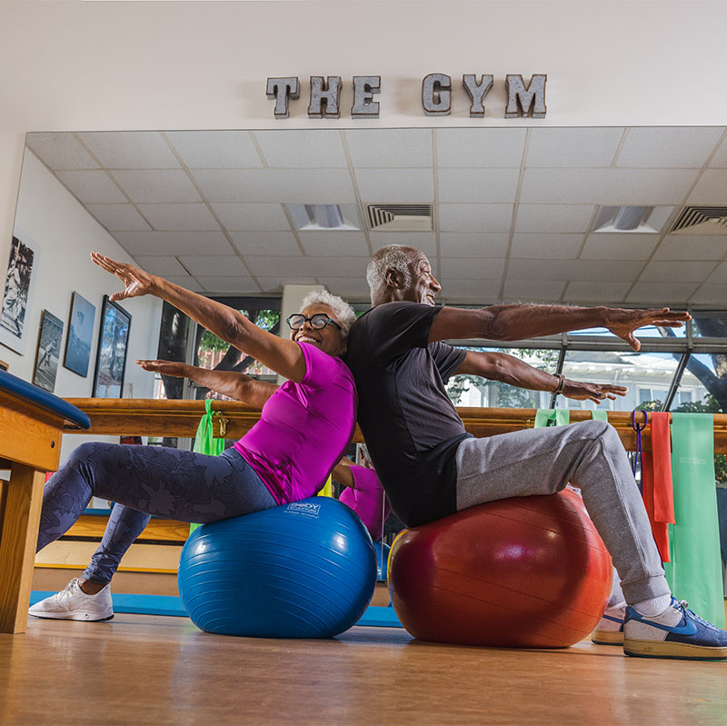 Senior man and woman sitting back-to-back on fitness balls in the Piedmont Gardens fitness center