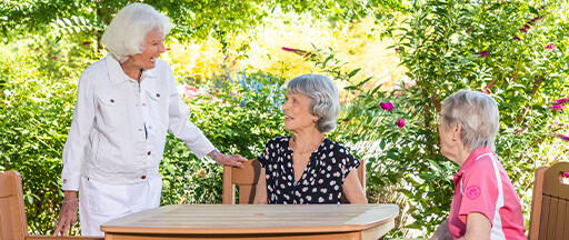 Senior woman standing and talking to two senior women sitting at an outdoor table in the garden at Rosewood