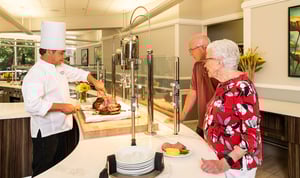 Senior couple at a carving station watching a chef carve meat in a dining room at Rosewood