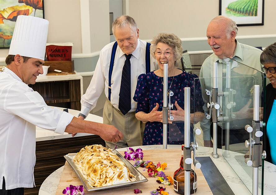 Two senior men and a senior woman watching a chef prepare food in a dining room at Rosewood
