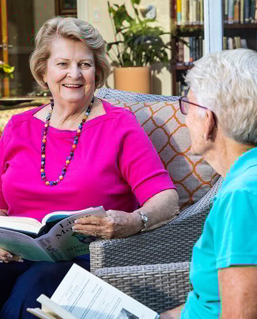 Two senior women sitting in armchairs and reading in the library at Royal Oaks