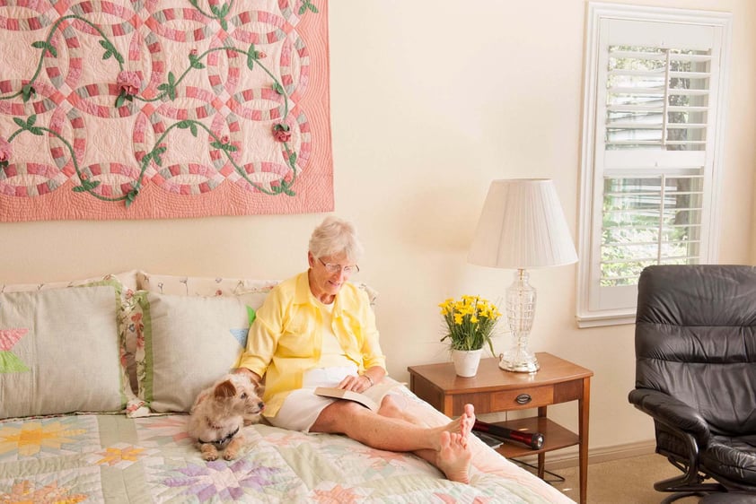 Senior woman sitting on her bed reading and petting a small dog in her apartment at Royal Oaks