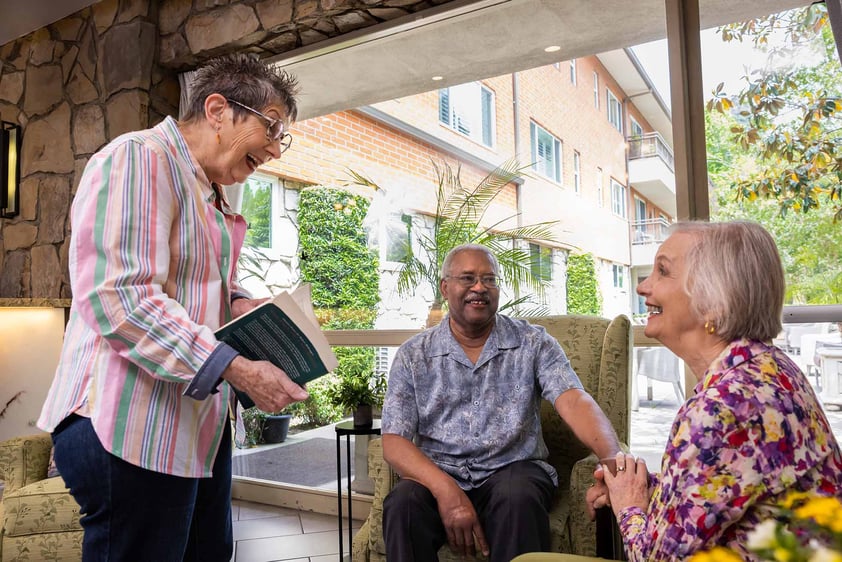 Senior woman holding a book talking to senior couple sitting in armchairs in a lounge area at Royal Oaks