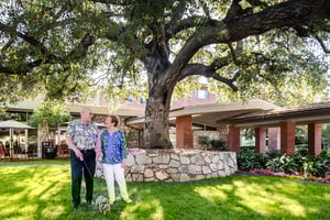 Senior couple holding a small dog standing under a large oak tree on the campus of Royal Oaks