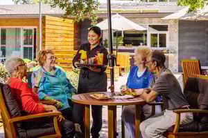 Server handing out cocktails to a group of four seniors sitting at a patio table under an umbrella at Royal Oaks