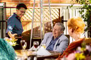 Server pouring a glass of red wine for a senior man sitting at an outdoor dining table