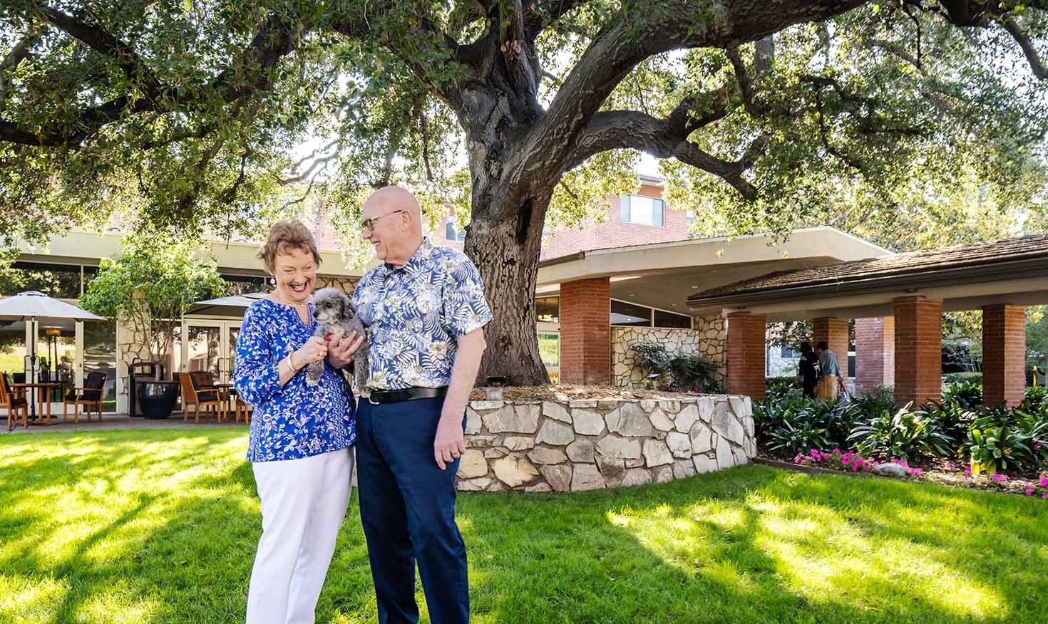 Senior couple holding a small dog standing under a large oak tree on the campus of Royal Oaks