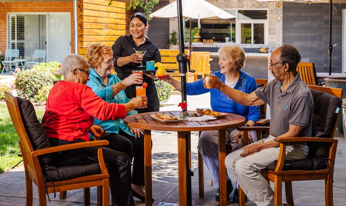 Server handing out cocktails to a group of four seniors sitting at a patio table under an umbrella at Royal Oaks