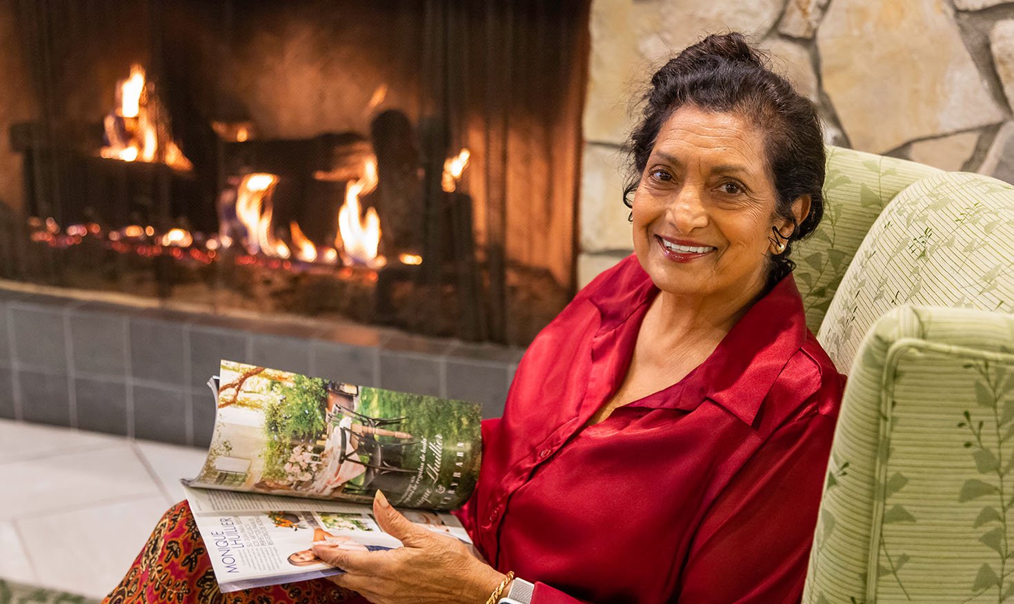 Senior woman holding a magazine and sitting by a fireplace 