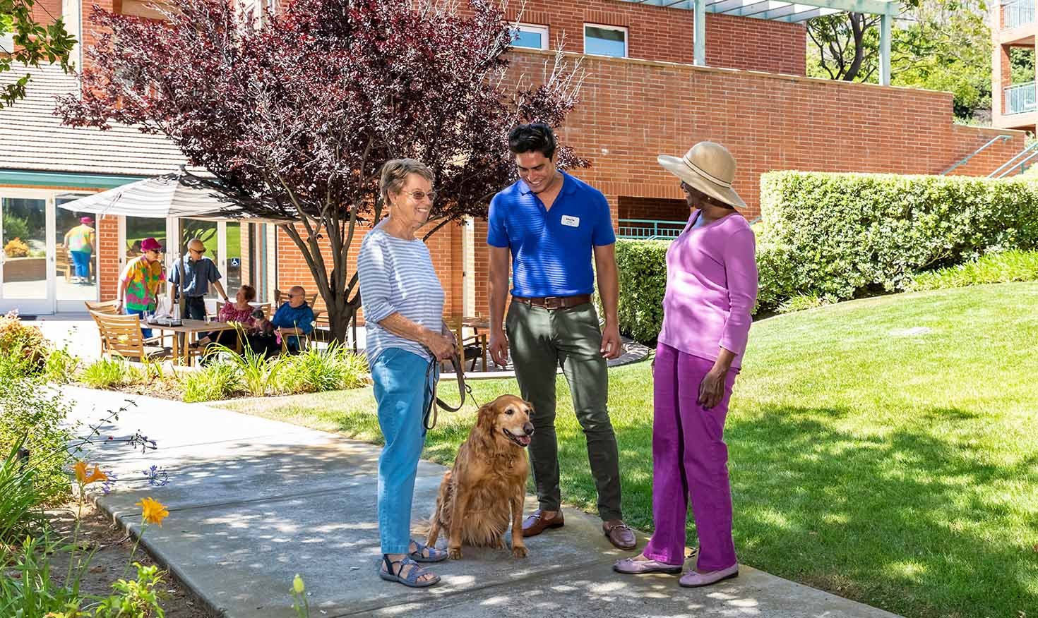 Senior woman with a dog on a leash talking with a senior woman and a male Royal Oaks team member on the sidewalk