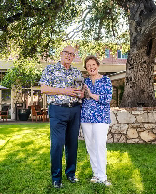 Senior couple holding a small dog standing under a large oak tree on the campus of Royal Oaks