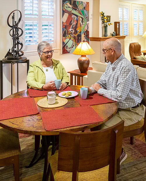 Senior couple holding mugs and sitting at the dining table of their cottage at Royal Oaks