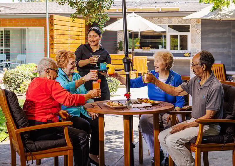 Server handing out cocktails to a group of four seniors sitting at a patio table under an umbrella at Royal Oaks