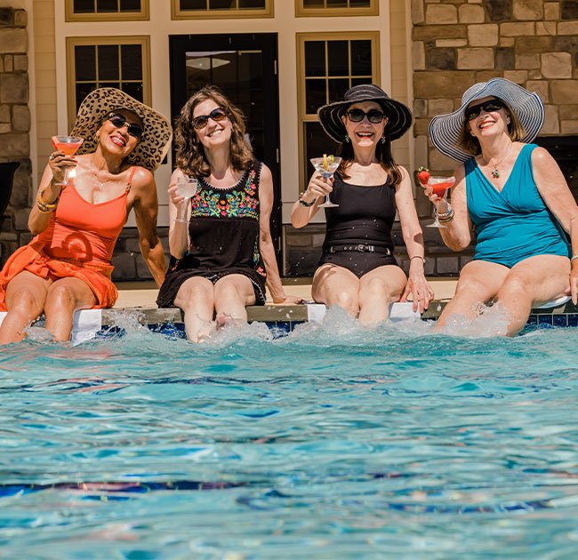 Four senior women in swimsuits sitting on the edge of the pool and holding up drinks