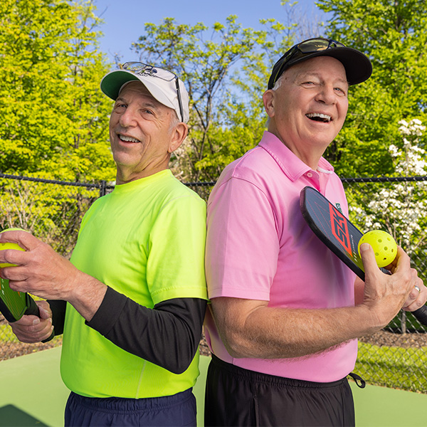 Two senior men in bright shirts holding pickleball paddles and balls on a court