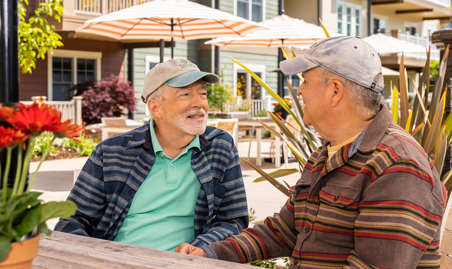 Two senior men wearing baseball caps sitting at an outdoor table at The Terraces at Los Altos