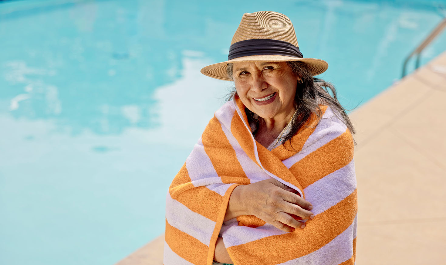 Senior woman in a hat standing by the pool wrapped in an orange and white striped towel