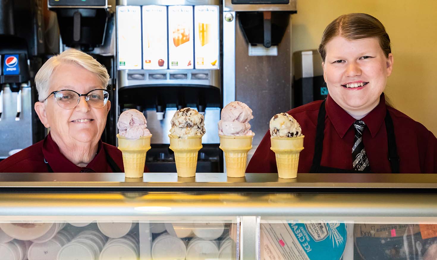Workers behind the counter of an ice cream parlor with four ice cream cones on the counter