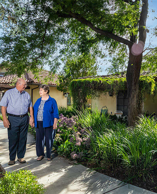 Senior man and woman holding hands and walking on sidewalk through garden