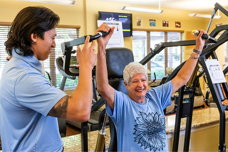 Senior woman using weight machine in fitness center with the assistance of personal trainer