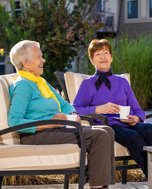 Two smiling senior women sitting outside drinking coffee