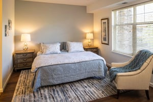 Bedroom with large bed, wooden nightstands, a rug and a chair covered with a throw in an apartment at The Terraces of Boise