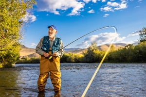 Senior man in waders, a fishing vest and baseball cap standing in a river fishing